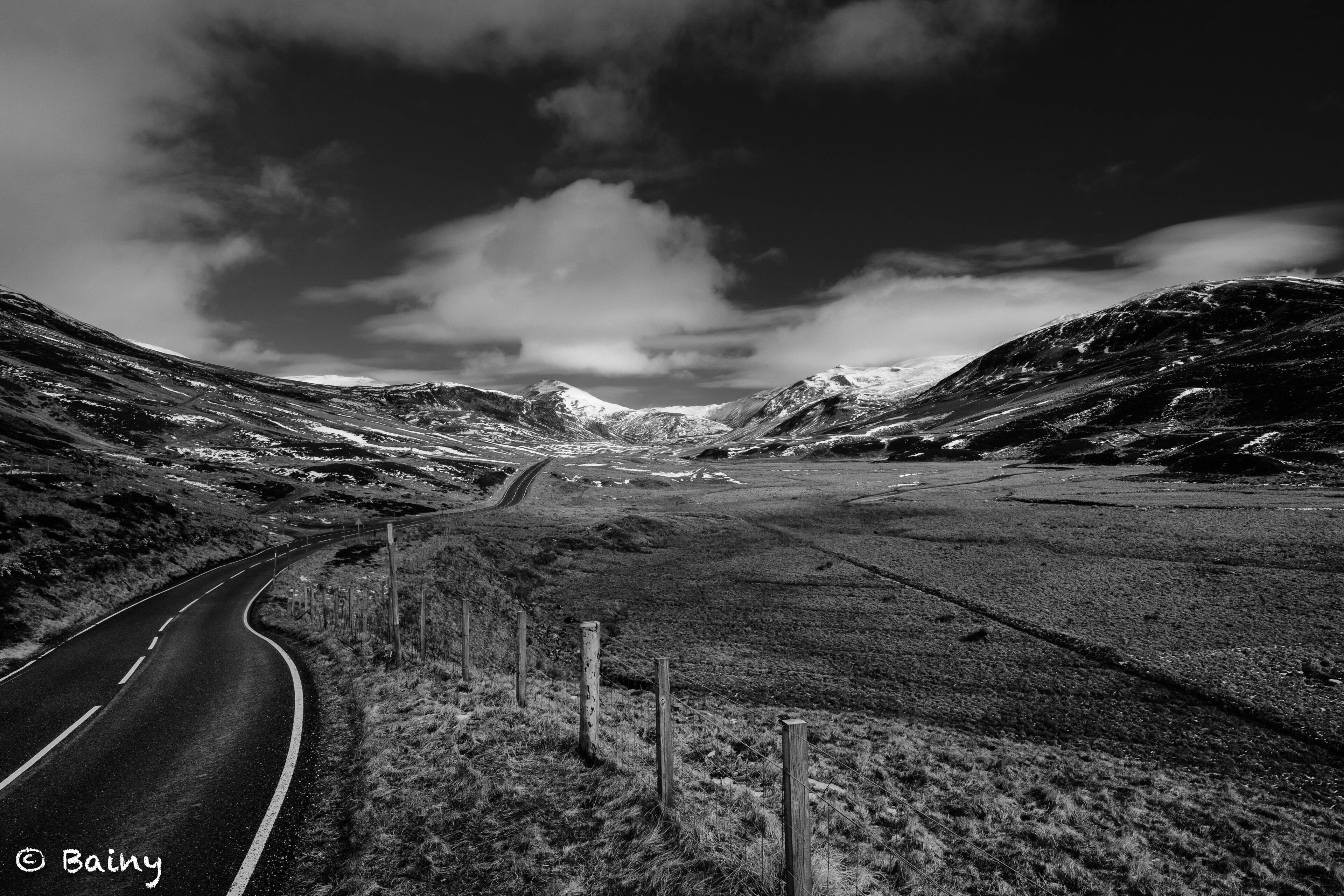 A winding road through a mountainous landscape with snow-covered peaks and a cloudy sky.
