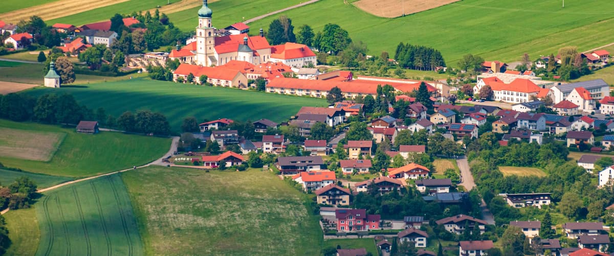 An aerial view of a village with a prominent church featuring a green dome and red roofs, surrounded by lush green fields and residential houses.