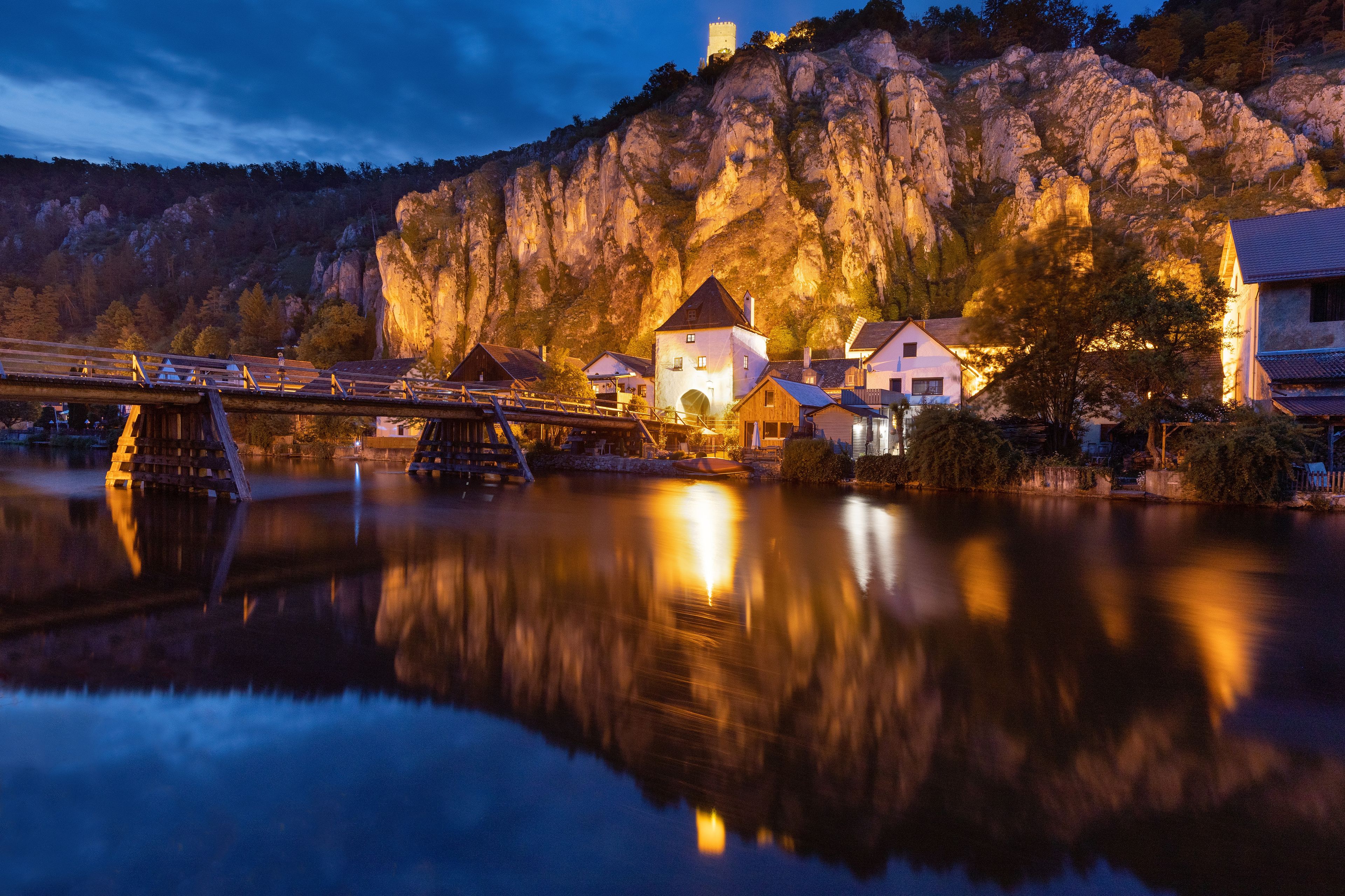 A river reflecting buildings and a bridge with a mountain backdrop.