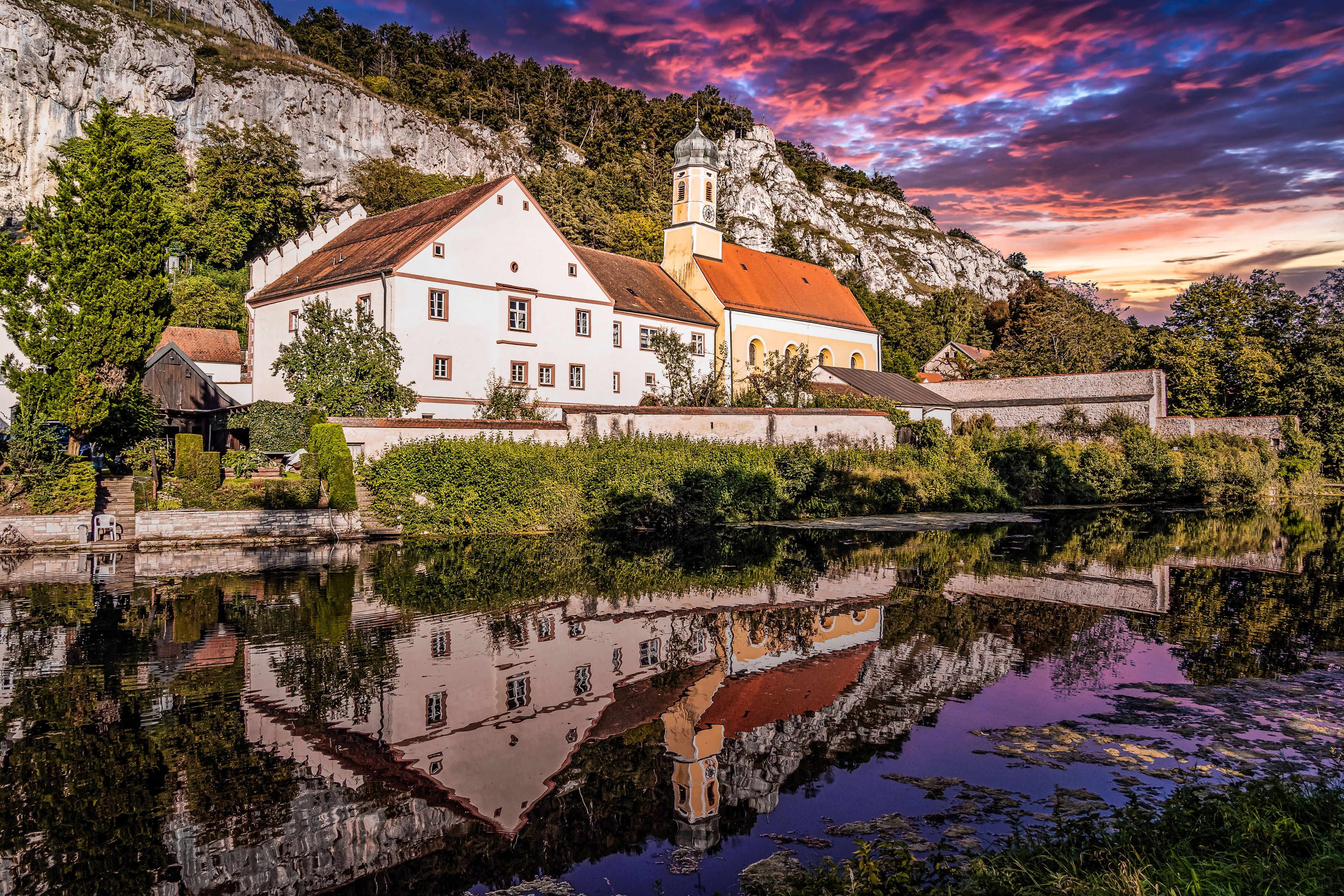 A white church with a red roof, surrounded by greenery and a body of water reflecting its image.