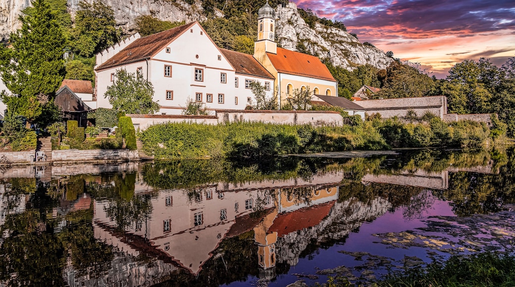 A white church with a red roof, surrounded by greenery and a body of water reflecting its image.