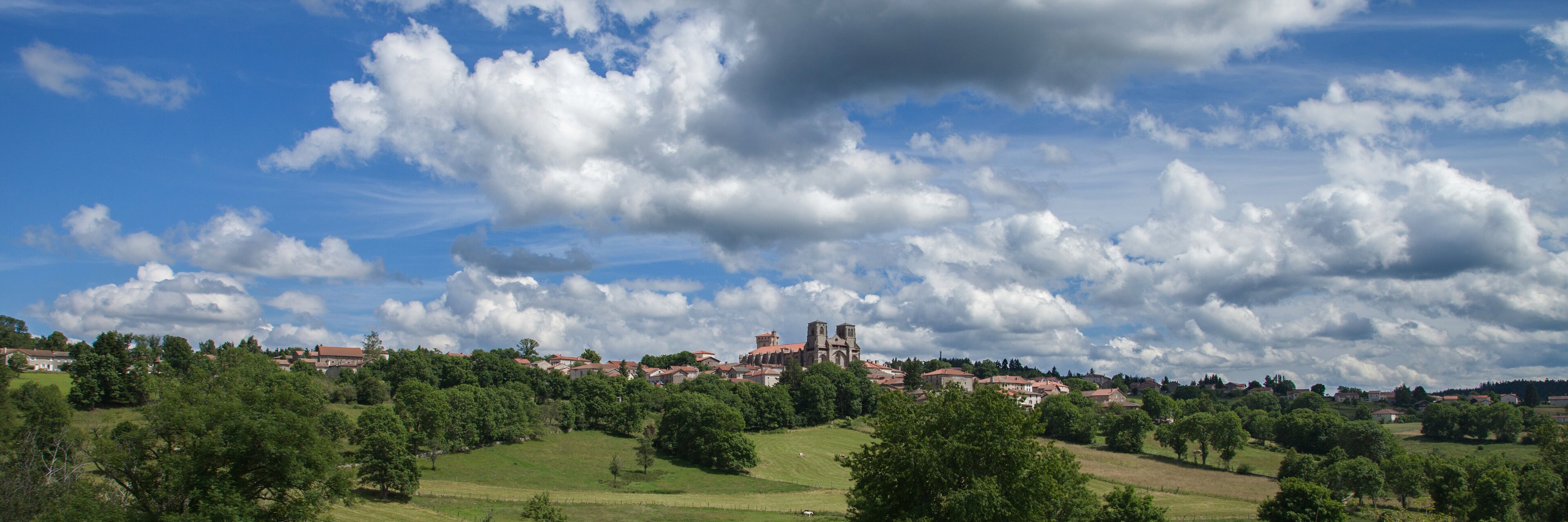 A town with a prominent church on a hill, surrounded by green fields and trees under a partly cloudy sky.