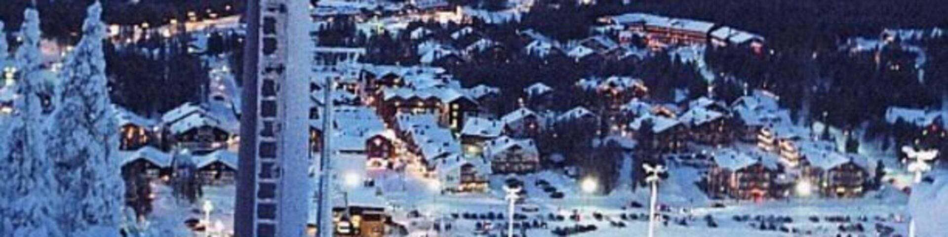 A snow-covered ski resort at dusk with illuminated buildings and a ski lift.