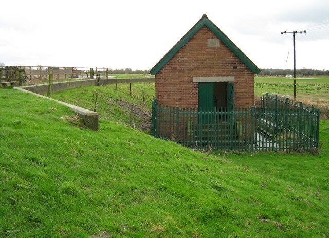 A small brick building with a green roof and a green door, surrounded by a green fence in a grassy field.
