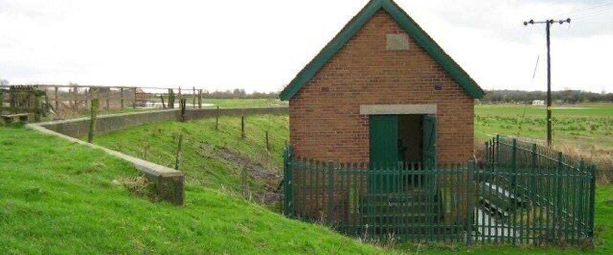 A small brick building with a green roof and a green door, surrounded by a green fence in a grassy field.