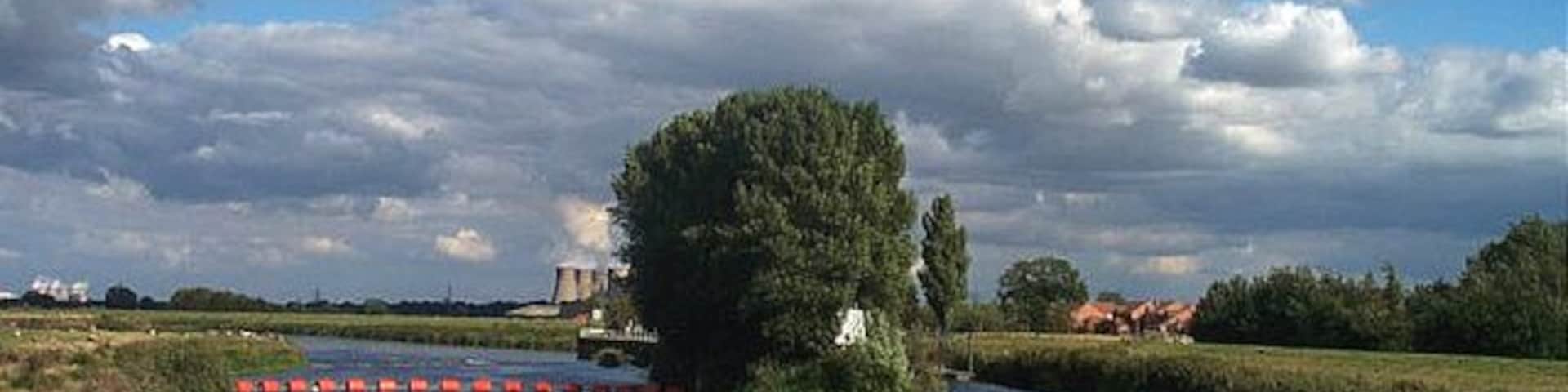 Beal- The River Aire. This photograph is taken from the Bridge over the River Aire at Beale, looking west. Eggborough Power Station can be seen on the horizon. The river is not navigable to the left of the island in the picture. There is a weir. To the right of the island is navigable via a lock.