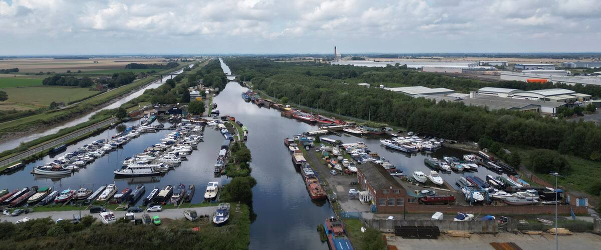 A river with boats docked on both sides, surrounded by greenery and industrial buildings.