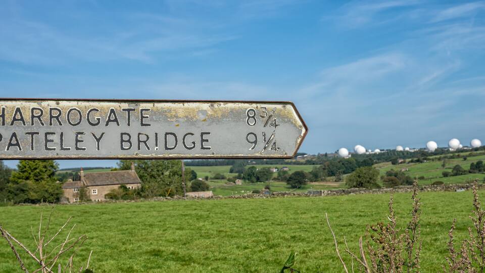 A weathered signpost with directions to Harrogate and Pateley Bridge, set against a rural landscape with fields and distant spherical structures.