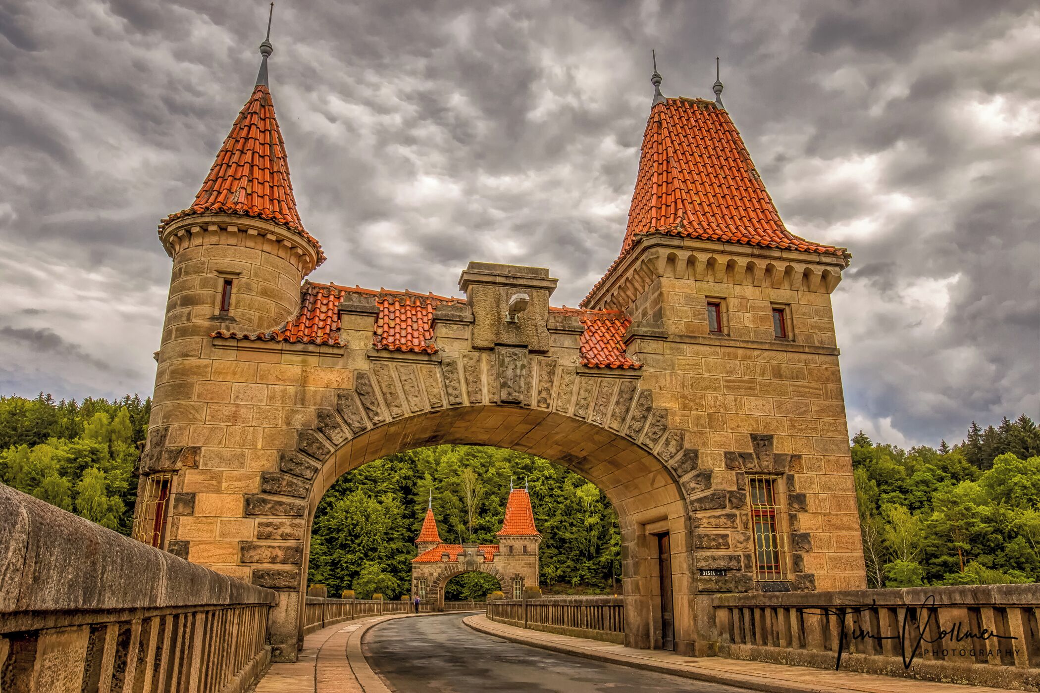 A stone bridge with two towers and a red-tiled roof, leading to a castle.