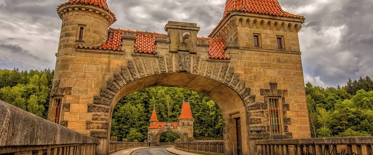 A stone bridge with two towers and a red-tiled roof, leading to a castle.