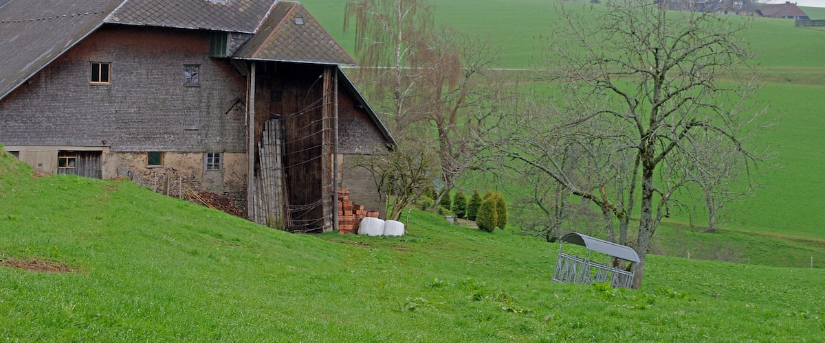 A rustic barn on a green hillside with a metal bench nearby.