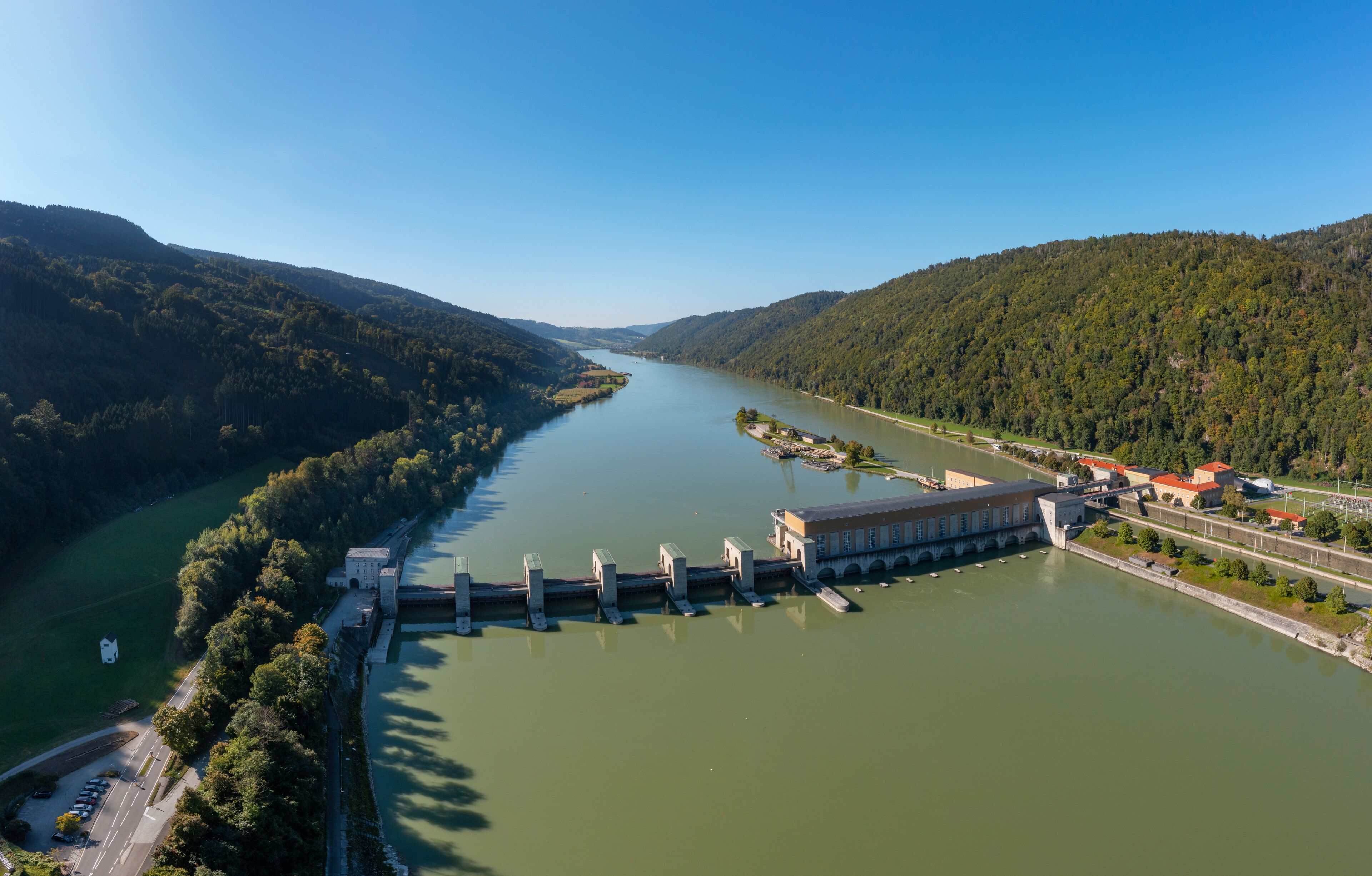 A dam with a hydroelectric power station, a river, and surrounding forested hills.