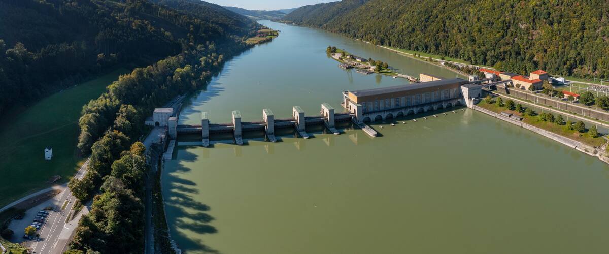 A dam with a hydroelectric power station, a river, and surrounding forested hills.