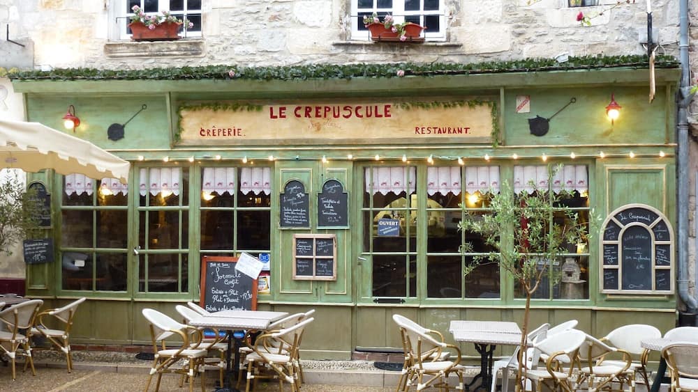 A charming outdoor cafe named 'Le Crepuscule' with green awnings, wicker chairs, and a chalkboard menu.