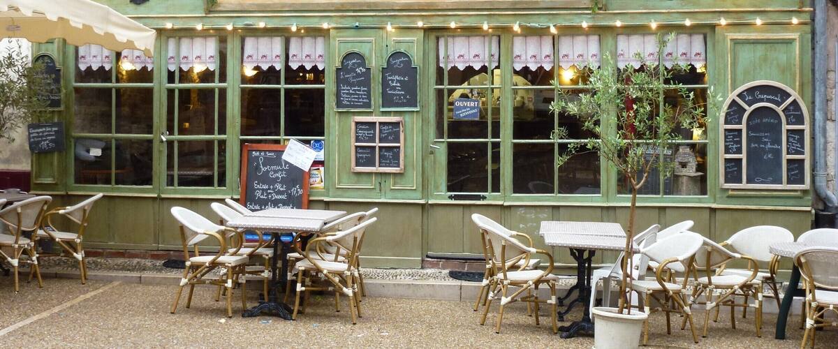 A charming outdoor cafe named 'Le Crepuscule' with green awnings, wicker chairs, and a chalkboard menu.