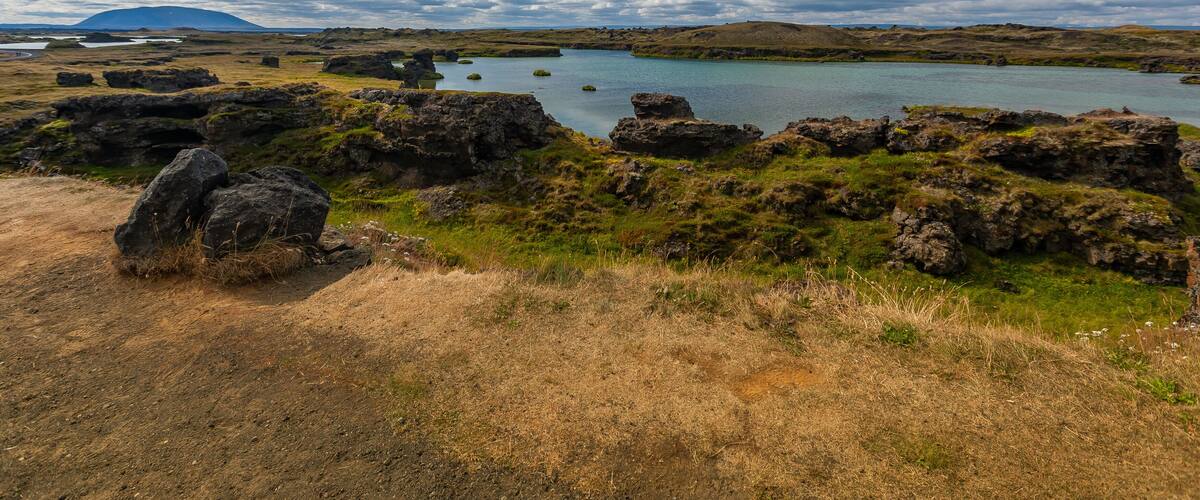 A coastal landscape with rocky terrain, a body of water, and a distant mountain under a partly cloudy sky.