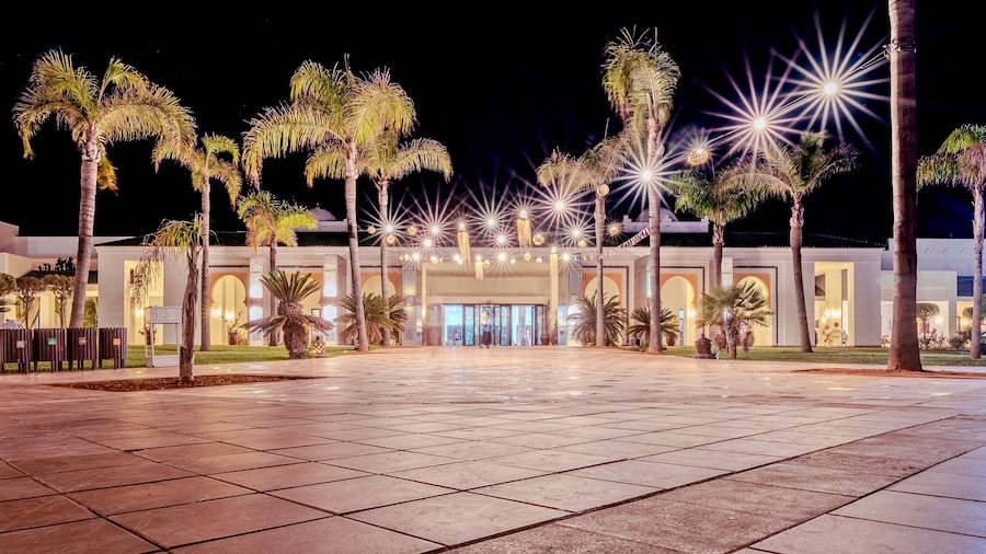 A well-lit building with a large entrance, surrounded by palm trees and a paved area.