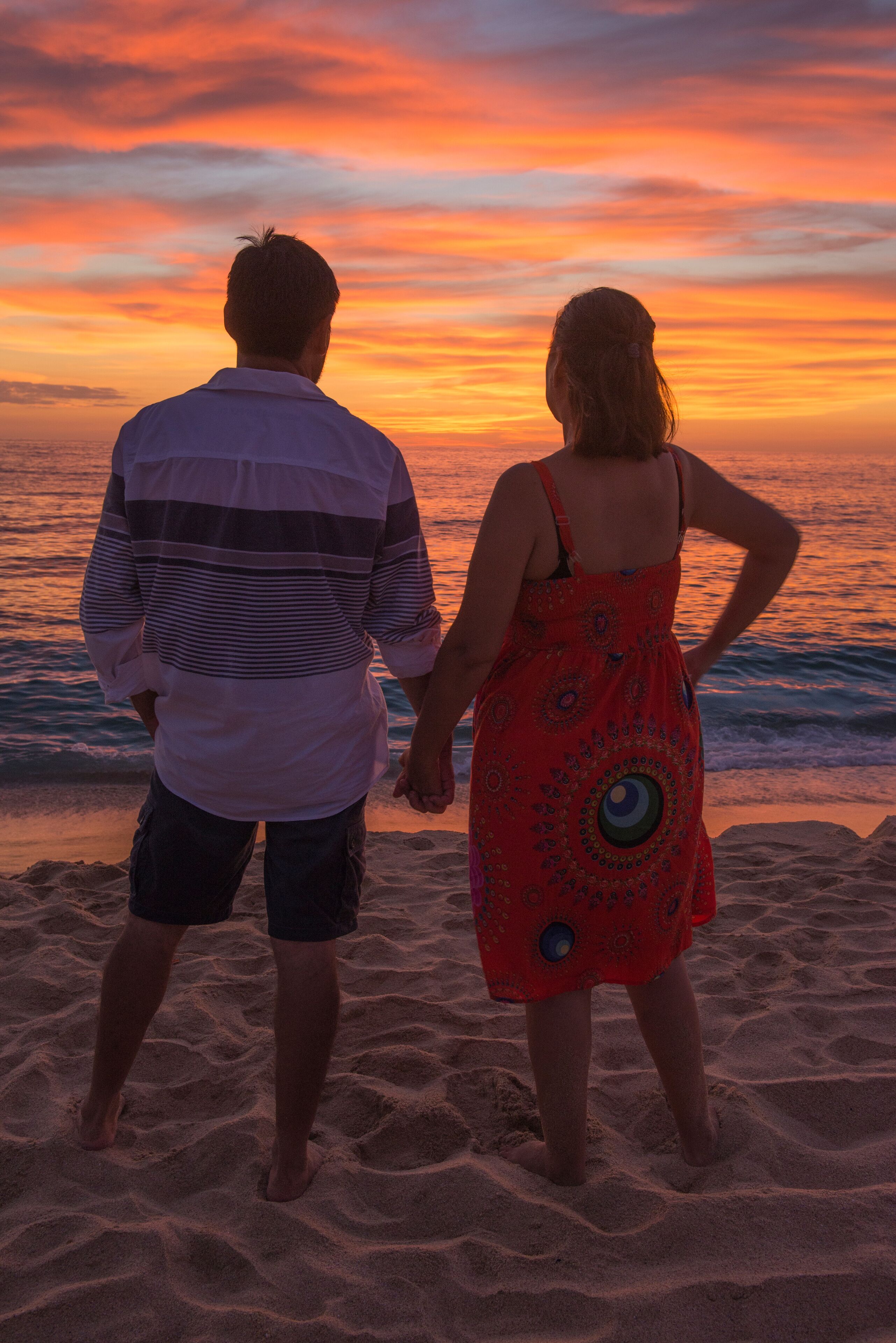 Um casal em pé na praia ao pôr do sol, com um céu colorido.