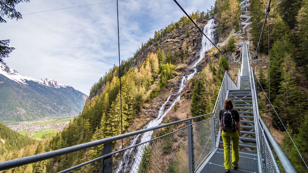 A person walking on a metal bridge over a waterfall in a mountainous area with a town in the valley below.
