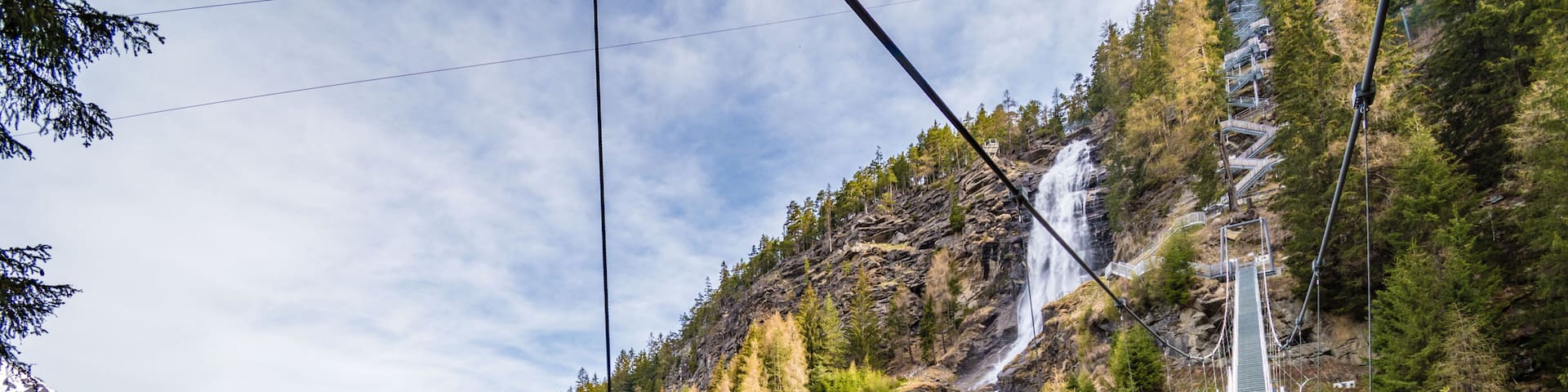 A person walking on a metal bridge over a waterfall in a mountainous area with a town in the valley below.