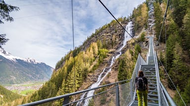 Una persona caminando sobre un puente de metal sobre un cascada en una zona montañosa, con un pueblo en el valle.