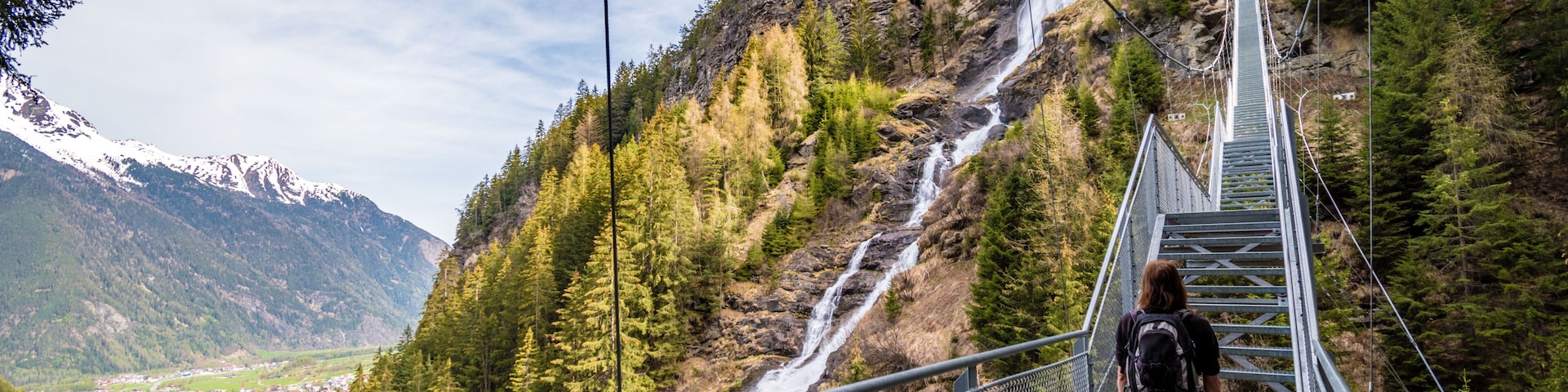 A person walking on a metal bridge over a waterfall in a mountainous area with a town in the valley below.