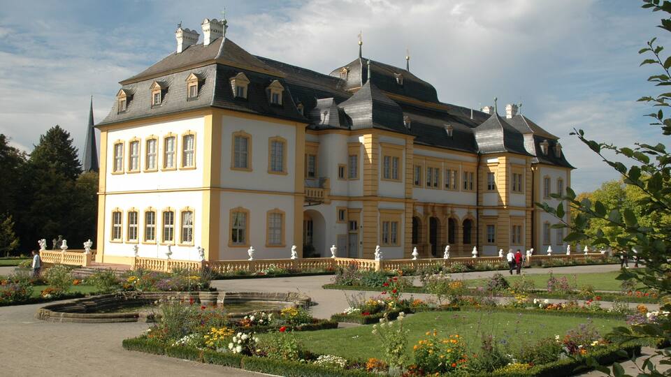 A large, historic building with a symmetrical facade, surrounded by a well-maintained garden with various flower beds and a central fountain.
