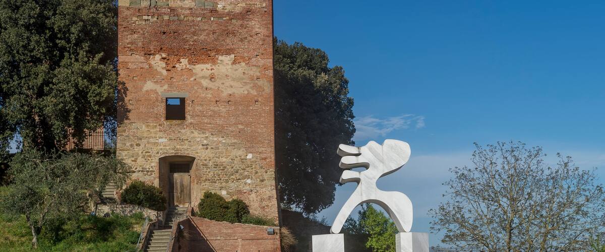 A historic brick tower with a narrow entrance, surrounded by trees and a modern sculpture.