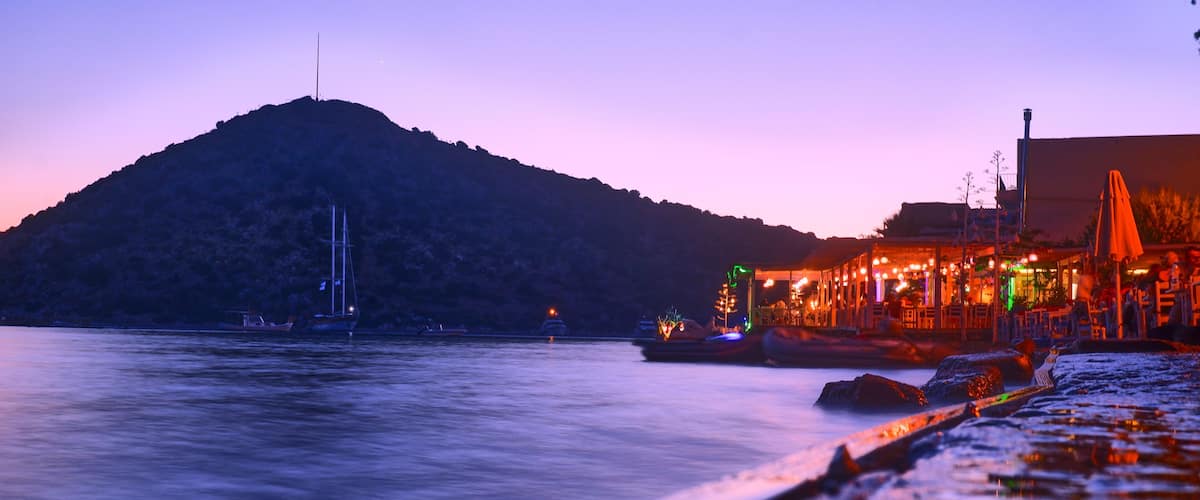 A coastal scene with a mountain, boats, and a brightly lit building.