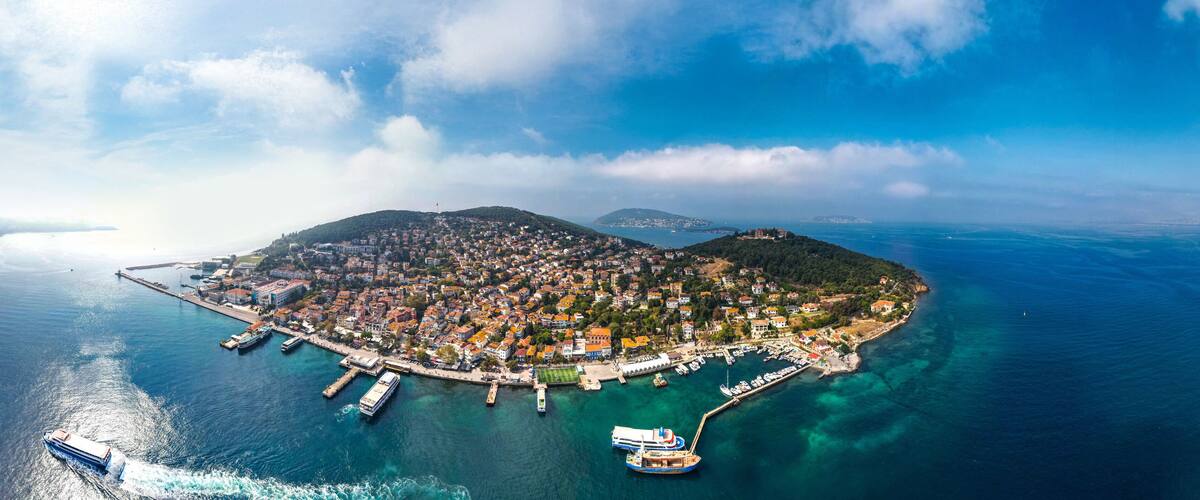 An aerial view of a coastal town with a harbor, boats, and a mountainous backdrop.