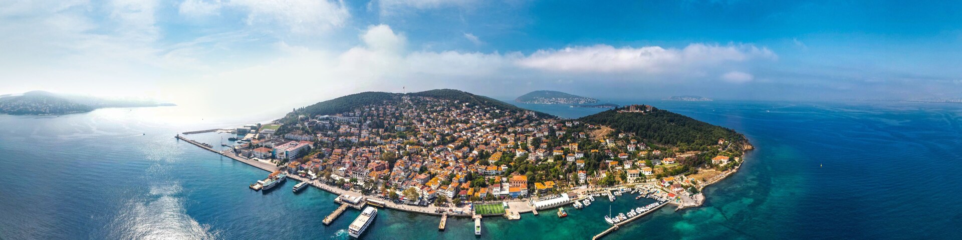 An aerial view of a coastal town with a harbor, boats, and a mountainous backdrop.