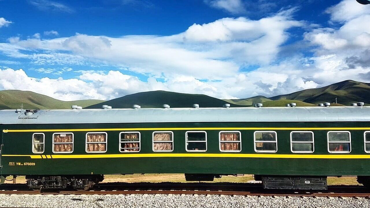 A green train with yellow stripes on a track, surrounded by mountains and a partly cloudy sky.