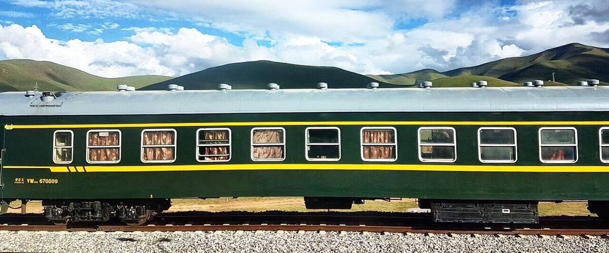 A green train with yellow stripes on a track, surrounded by mountains and a partly cloudy sky.