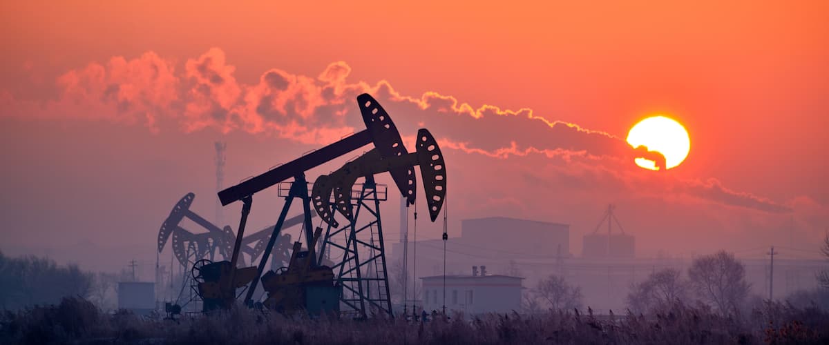 An oil pumpjack silhouetted against a sunset with industrial buildings in the background.