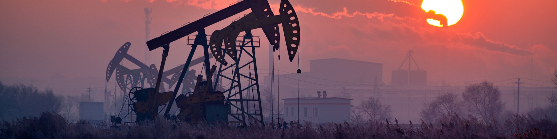 An oil pumpjack silhouetted against a sunset with industrial buildings in the background.