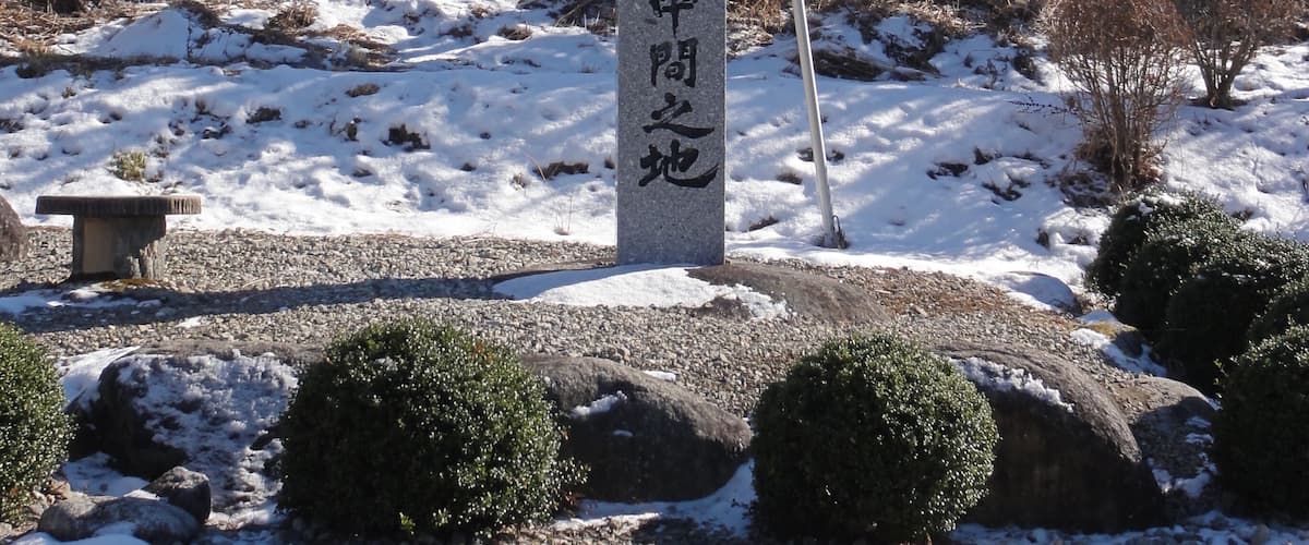 Ein Steinsockel mit japanischen Schriftzeichen, umgeben von verschneitem Boden, Steinen und Bäumen, mit Bergen im Hintergrund.