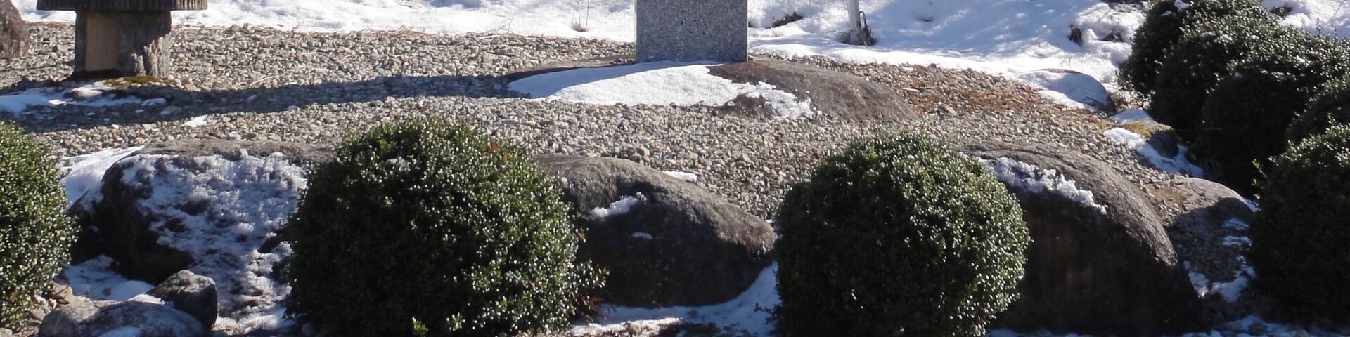 A stone monument with Japanese characters, surrounded by snow-covered ground, rocks, and trees, with mountains in the background.