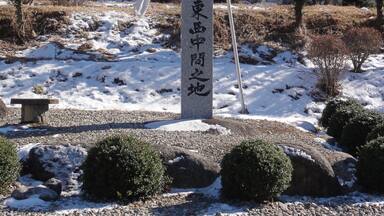 Monumento de piedra con caracteres japoneses, rodeado de terreno nevado, rocas y árboles, con montañas al fondo.