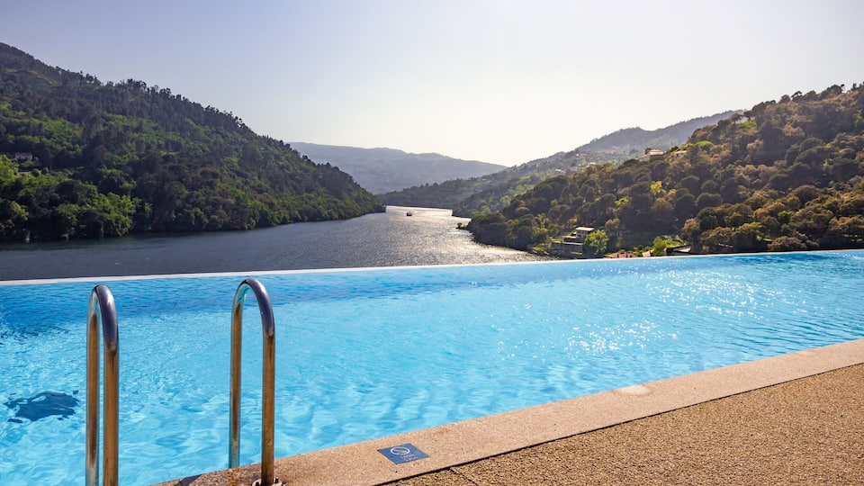 A swimming pool with metal ladders, surrounded by a forested hillside and a body of water.