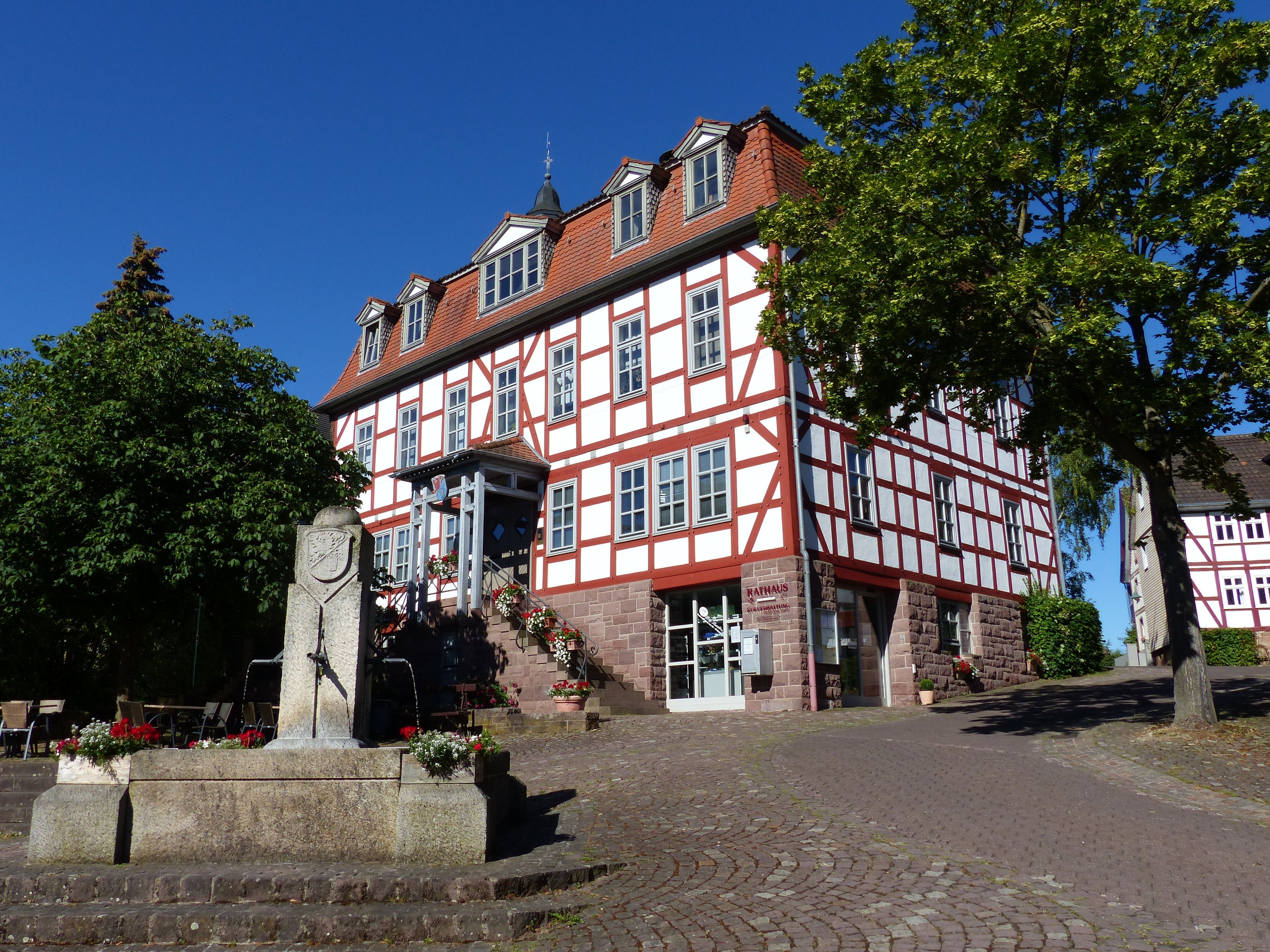 A traditional half-timbered building with a red and white facade, a stone monument, and cobblestone paving.