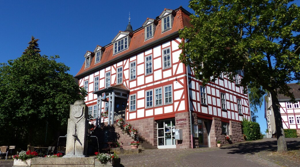 A traditional half-timbered building with a red and white facade, a stone monument, and cobblestone paving.