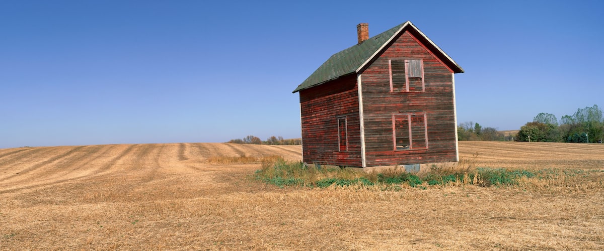 A red wooden house with a green roof and a chimney, situated in a field with rows of crops under a clear blue sky.