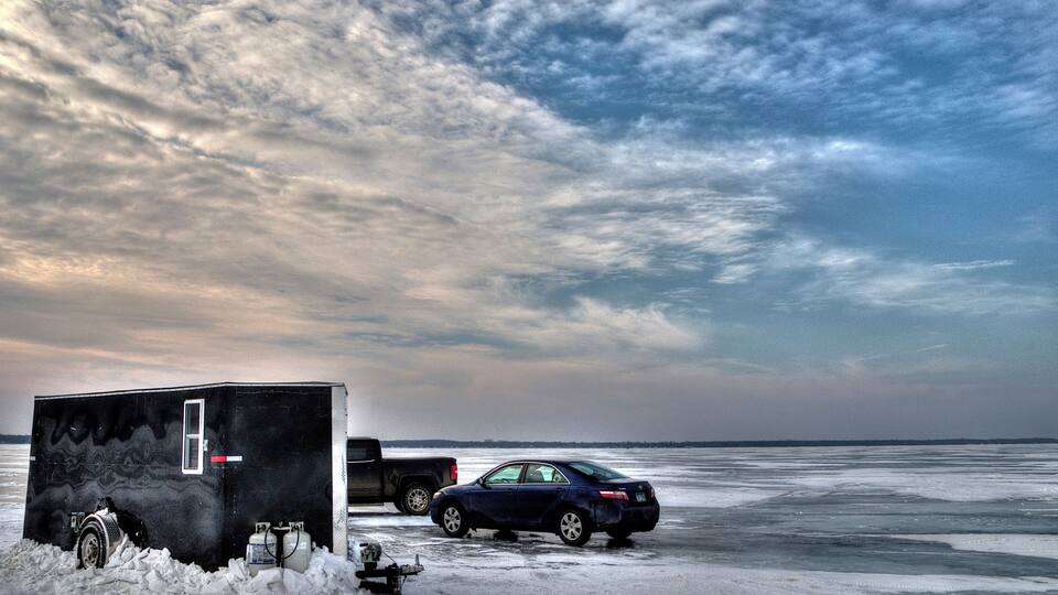 A snowy landscape with a car and a trailer on a frozen body of water, under a cloudy sky.