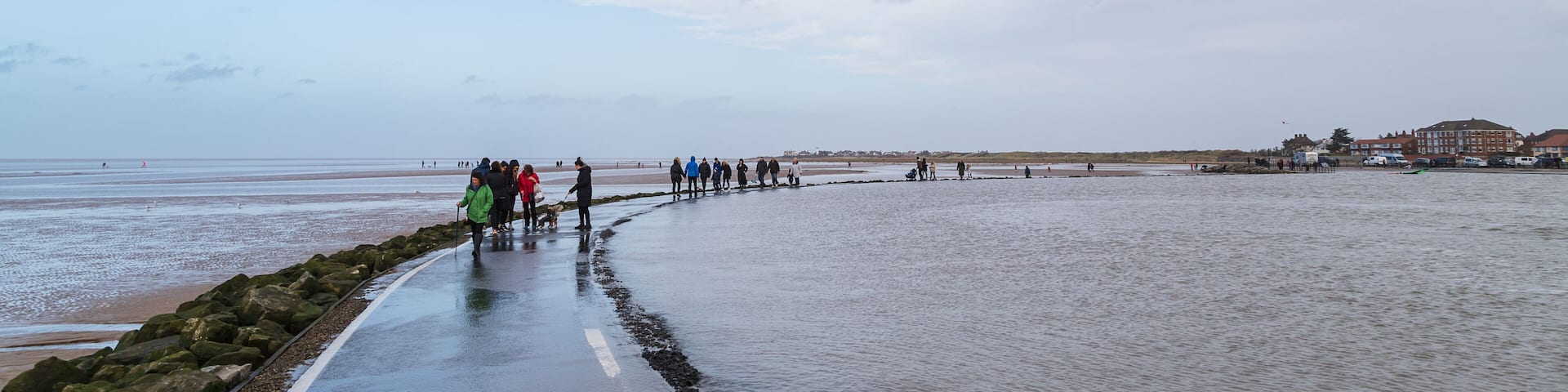 A wet beach with people walking along a path, a rocky shoreline, and buildings in the background.