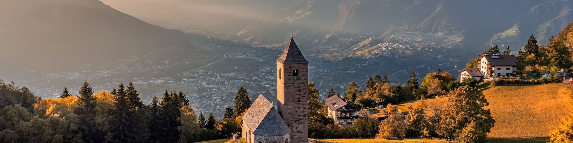A church with a bell tower, surrounded by mountains and autumn trees.