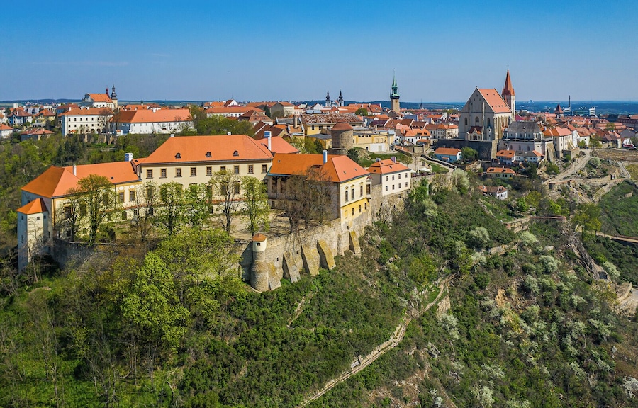 Une ville historique abritant un château imposant, perché sur une colline, entourée de bâtiments aux toits rouges et d’une église dotée d’un clocher élancé.