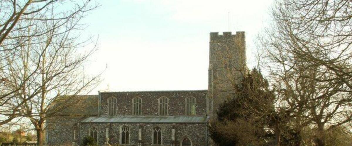 A church with a tower and arched windows, surrounded by a hedge and a rural road.
