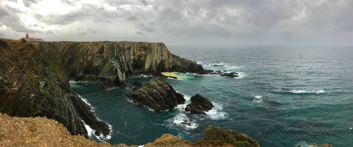 A coastal cliff with a lighthouse, rocky shoreline, and turbulent sea.