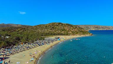 A beach with clear blue water, palm trees, and a rocky hill in the background.