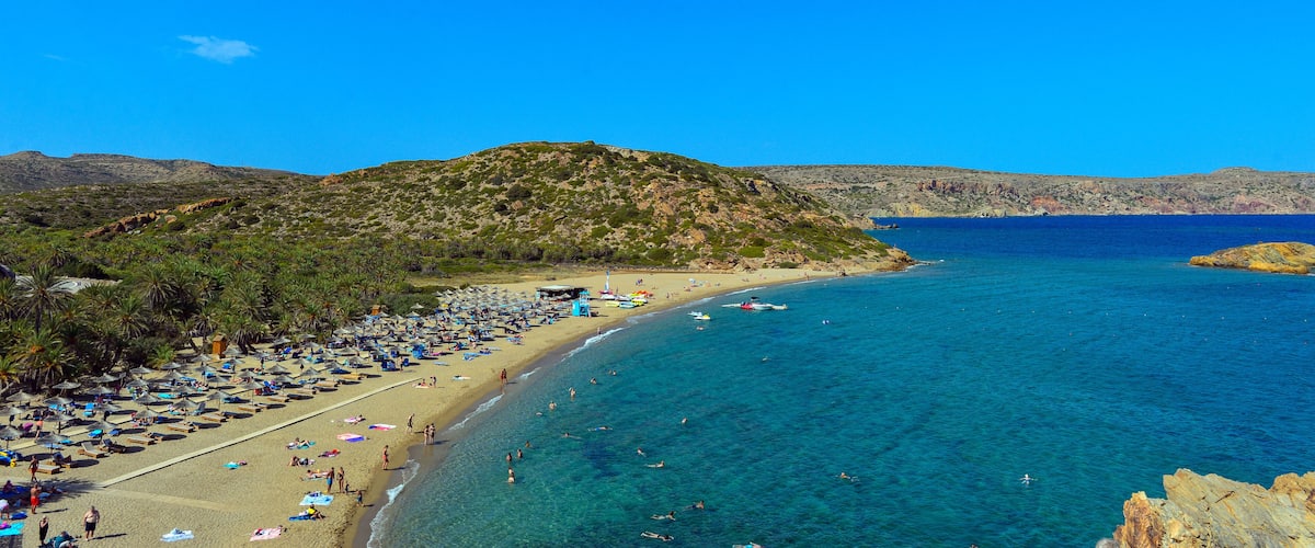 A beach with clear blue water, palm trees, and a rocky hill in the background.